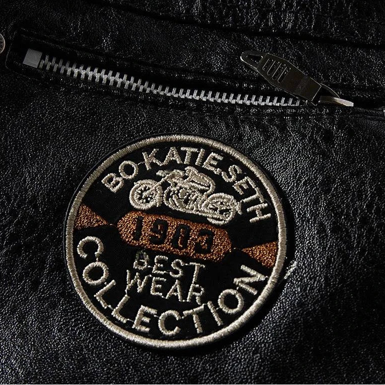 Detalhe aproximado do patch redondo 'BO KATIE SETH 1983 BEST WEAR COLLECTION' com moto, em jaqueta de couro preta com zíper prateado. Exclusividade e autenticidade.