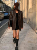 Mulher caminhando em Embu das Artes, usando jaqueta de couro oversized feminina marrom, estilo blazer, sobre vestido preto e com botas altas, beanie e bolsa a tiracolo, exibindo um visual de luxo urbano e elegante para o inverno.