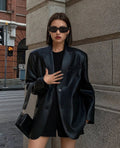 Mulher usando jaqueta de couro oversized feminina preta, estilo blazer com caimento amplo, um botão frontal e bolsos laterais, combinada com óculos escuros e bolsa, exibindo um visual de luxo moderno e exclusivo para as ruas de Embu das Artes.
