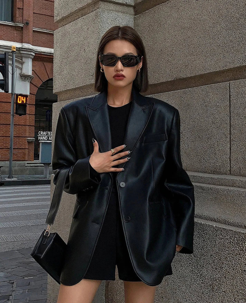 Mulher usando jaqueta de couro oversized feminina preta, estilo blazer com caimento amplo, um botão frontal e bolsos laterais, combinada com óculos escuros e bolsa, exibindo um visual de luxo moderno e exclusivo para as ruas de Embu das Artes.