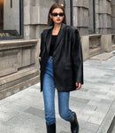 Mulher em jaqueta de couro oversized preta, jeans azul e botas pretas, posando em fachada de concreto