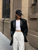 Mulher com jaqueta de couro oversized preta, top branco cropped, calça cinza e boné preto