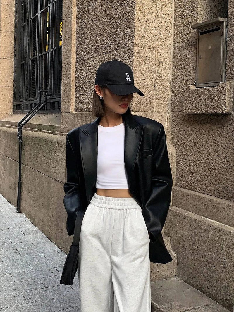 Mulher usando jaqueta de couro oversized feminina preta, estilo blazer, combinada com top cropped branco, calça de moletom cinza e boné preto, exibindo um visual de luxo descontraído e exclusivo para o estilo urbano de Embu das Artes.