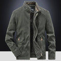 Jaqueta masculina bomber parka verde escuro com gola alta, zíperes duplos (interno e externo), múltiplos bolsos com zíperes e botões de pressão, detalhes em couro nos puxadores, e bordado "DEFENDER 19 JP 71" no peito. O forro de pelúcia preto é visível. Uma peça de alta qualidade e funcionalidade, combinando o estilo militar e o conforto térmico