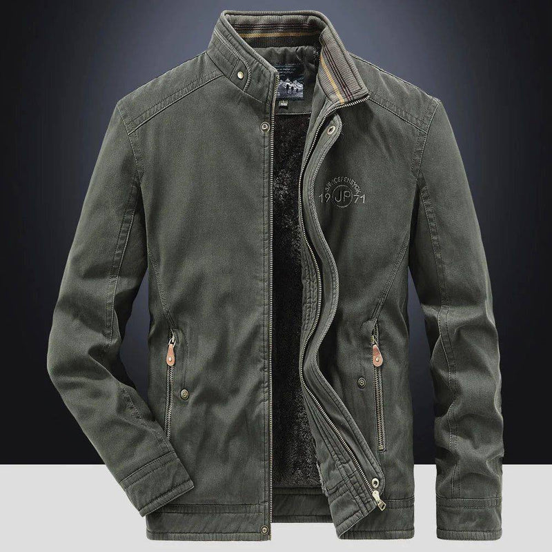 Jaqueta masculina bomber parka verde escuro com gola alta, zíperes duplos (interno e externo), múltiplos bolsos com zíperes e botões de pressão, detalhes em couro nos puxadores, e bordado "DEFENDER 19 JP 71" no peito. O forro de pelúcia preto é visível. Uma peça de alta qualidade e funcionalidade, combinando o estilo militar e o conforto térmico