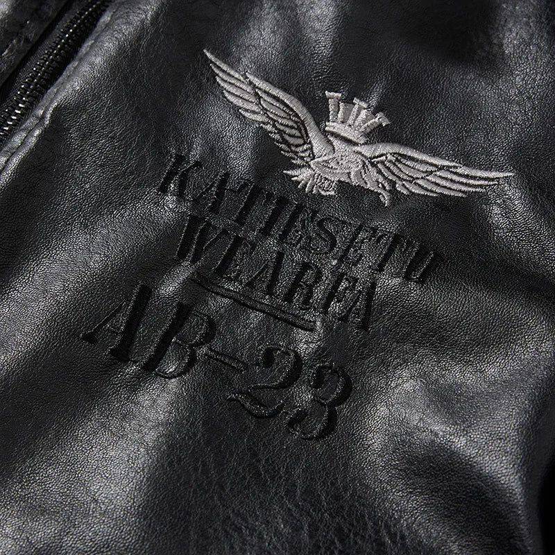 Close-up do detalhe bordado no ombro da jaqueta masculina de couro preta, com um emblema de asas e coroa em cinza, e as inscrições em relevo "KATIE SETH WEAR" e "AB-23". O bordado detalhado e a textura do couro conferem um toque de exclusividade e autenticidade à peça.