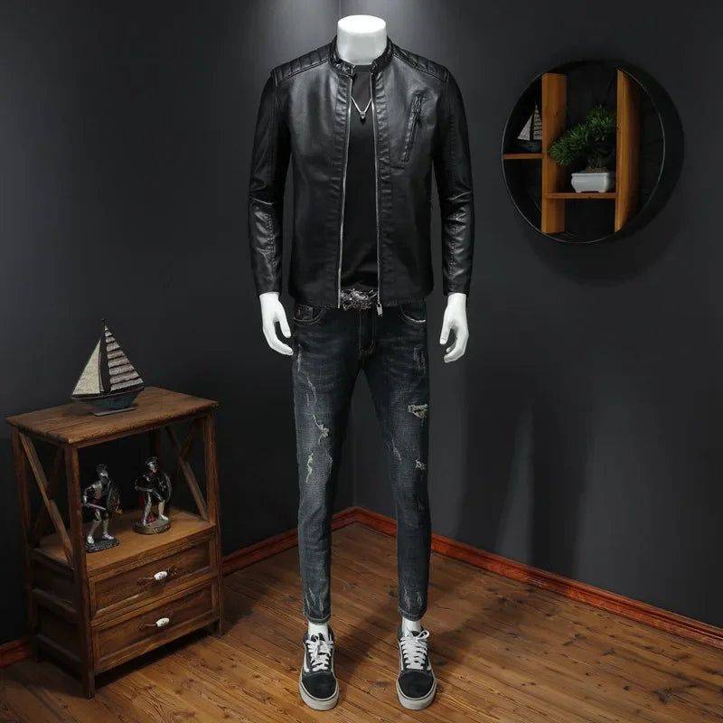 Manequim masculino vestindo jaqueta de couro premium preta com gola mandarim, camiseta preta, jeans skinny rasgados e tênis pretos e brancos. À esquerda, uma mesa de madeira com decorações, e à direita, uma prateleira de parede com plantas. A jaqueta confere um estilo moderno e exclusivo, perfeito para o ambiente sofisticado de Embu das Artes.