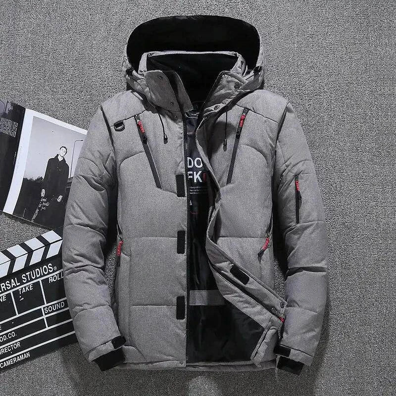 Homem vestindo jaqueta masculina parka acolchoada cinza com capuz preto, múltiplos bolsos com zíperes vermelhos e pretos, e fechamento frontal com zíper e botões de pressão, em um ambiente interno com elementos de produção cinematográfica ao fundo. Uma peça moderna e funcional, ideal para quem busca um estilo exclusivo e prático para o clima de Embu das Artes.