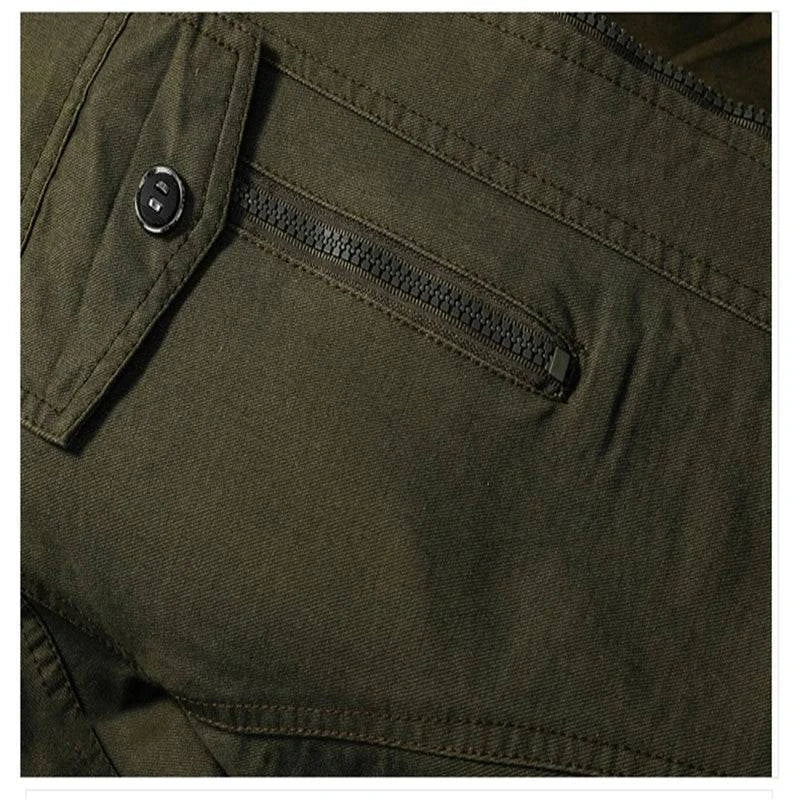 Detalhe aproximado do bolso na jaqueta militar tática verde oliva, mostrando zíper preto, lapela com botão e costuras reforçadas no tecido. Funcionalidade e atenção aos detalhes de design.