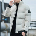 Homem usando jaqueta puffer acolchoada cinza claro com gola alta e fechamento em zíper cinza escuro, com punhos e barra canelados pretos e um distintivo circular escuro na manga esquerda. Leveza e calor para um visual moderno no inverno.