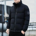 Homem usando jaqueta puffer acolchoada preta com gola alta e fechamento em zíper cinza, com punhos e barra canelados pretos e um distintivo circular escuro na manga esquerda. Essencial para o frio, combinando estilo clássico e conforto superior.