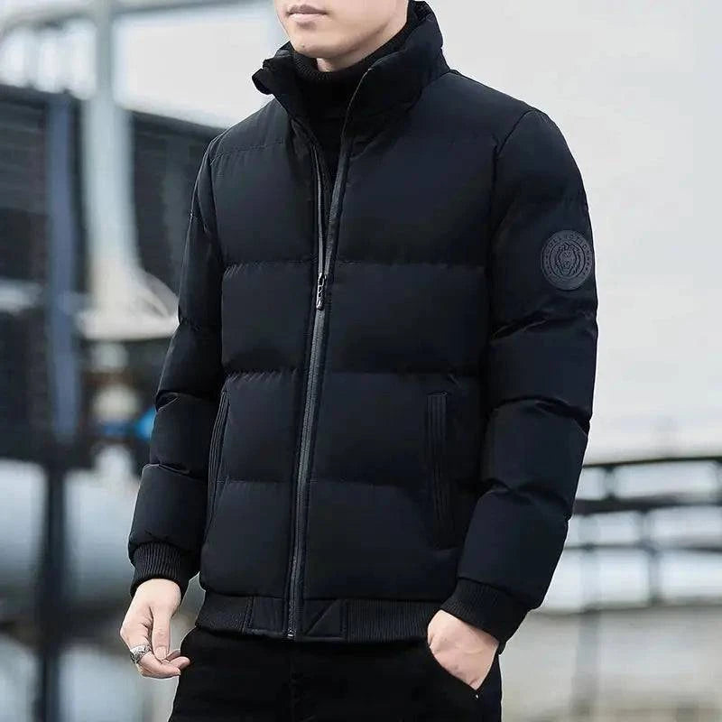 Homem usando jaqueta puffer acolchoada preta com gola alta e fechamento em zíper cinza, com punhos e barra canelados pretos e um distintivo circular escuro na manga esquerda. Essencial para o frio, combinando estilo clássico e conforto superior.