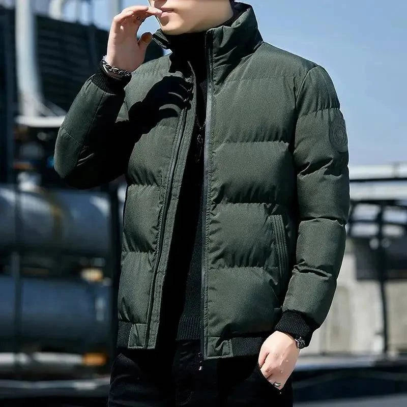 Homem usando jaqueta puffer acolchoada verde-escuro com gola alta, zíper preto aparente e distintivo circular escuro na manga, em ambiente urbano com iluminação solar. Peça essencial para os dias frios, combinando funcionalidade e um estilo sutilmente sofisticado.