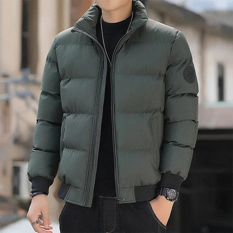 Homem usando jaqueta puffer acolchoada verde oliva com gola alta e fechamento em zíper preto, com punhos e barra canelados pretos, em ambiente urbano. Uma peça essencial e confortável para os dias frios com um toque de sofisticação.