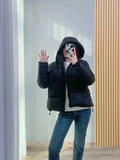 Modelo usando jaqueta puffer feminina preta com capuz e caimento moderno, combinada com jeans e suéter branco, em pose de selfie, exibindo um visual luxuoso e casual para o inverno.
