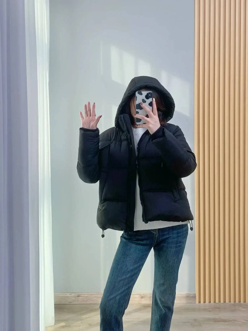 Modelo usando jaqueta puffer feminina preta com capuz e caimento moderno, combinada com jeans e suéter branco, em pose de selfie, exibindo um visual luxuoso e casual para o inverno.