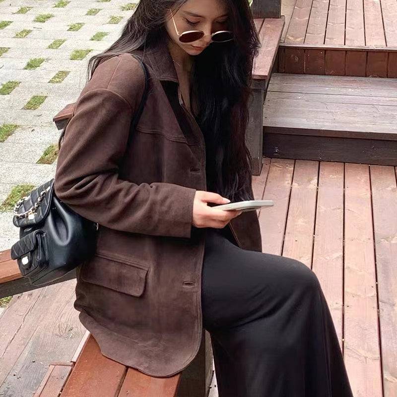 Mulher sentada em degraus de madeira, usando jaqueta retro feminina de pele de carneiro marrom, combinada com calça preta, óculos de sol e mochila, exibindo um estilo luxuoso e descontraído, perfeito para o charme de Embu das Artes.