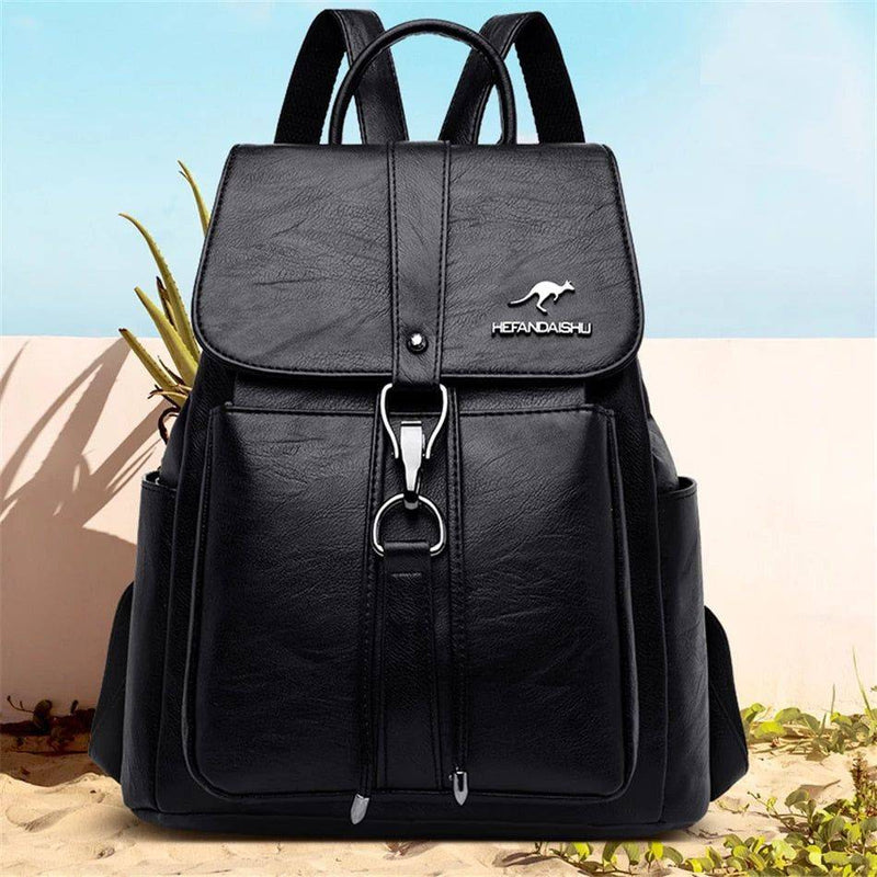 Mochila e bolsa feminina de couro 2 em 1 preta com aba frontal e fecho de gancho, exibida em ambiente externo com fundo de praia, destacando seu design versátil para diferentes ocasiões.