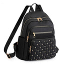 Mochila feminina pequena de nylon preta com bolso frontal matelassê decorado com tachas douradas, múltiplos zíperes e alças ajustáveis, destacando o design fashion e funcional.