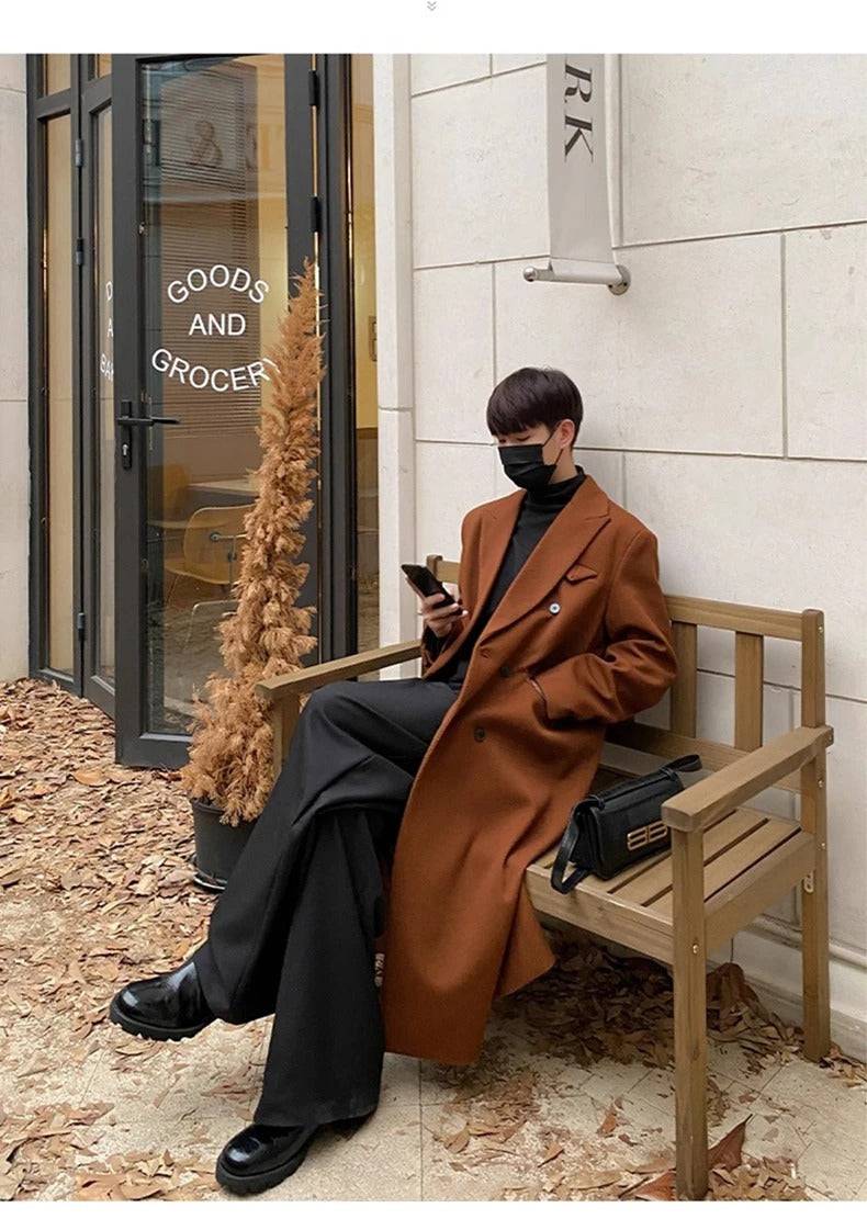 Homem usando sobretudo masculino longo social Eik marrom/caramelo, calça preta e máscara facial, sentado em banco de madeira em ambiente externo urbano, com uma pequena bolsa preta ao lado.