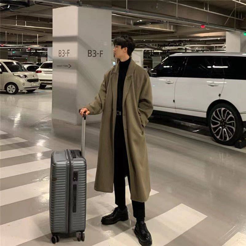 Sobretudo masculino Zellum Elite longo marrom-claro, solto, em garagem de estacionamento de luxo com mala de viagem e carro branco ao fundo, evocando estilo exclusivo e pronto para jornadas.
