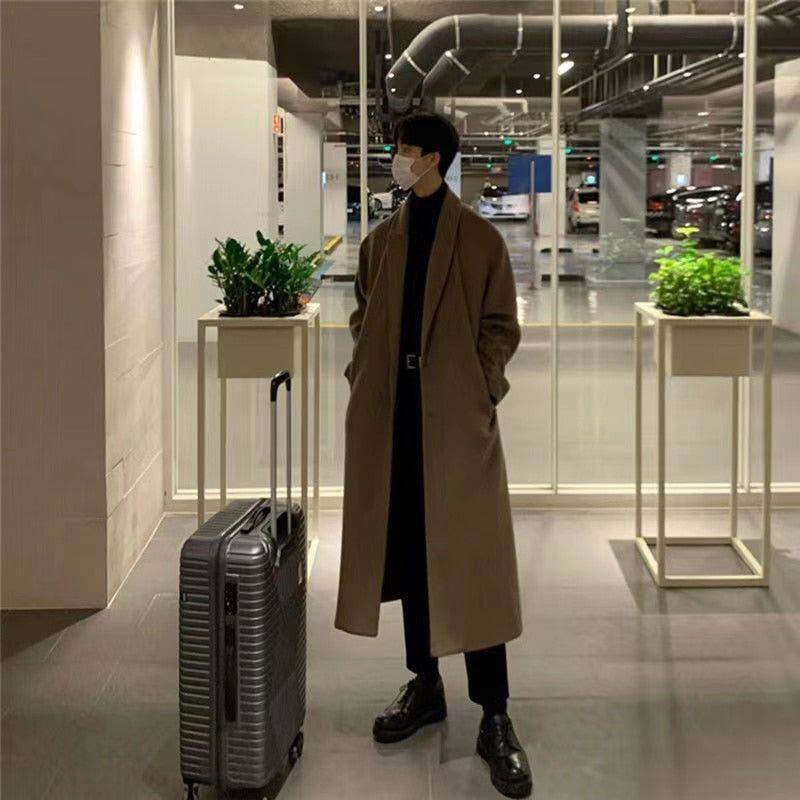 Sobretudo masculino Zellum Elite longo marrom-claro, solto, usando máscara e com mala de viagem em ambiente de garagem moderna, evocando estilo exclusivo e preparado para viagens.