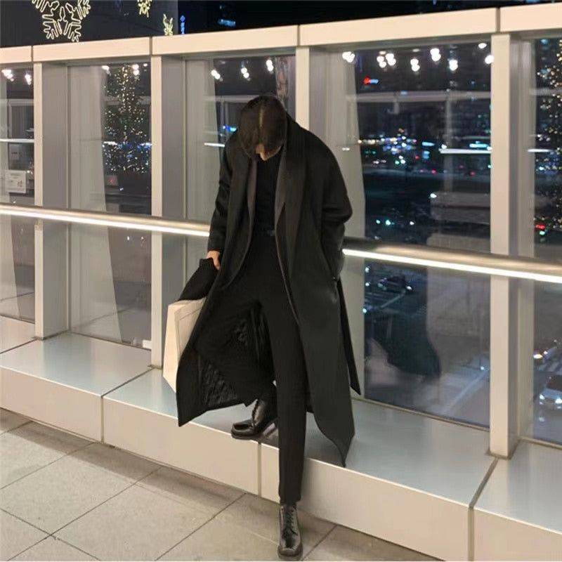 Sobretudo masculino Zellum Elite longo preto, solto, sobre gola alta e calça preta, em ambiente urbano noturno com luzes refletindo, segurando sacola de compras, evocando estilo exclusivo e sofisticado.