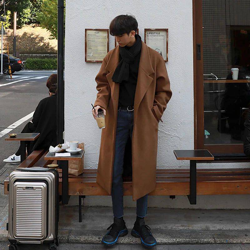 Sobretudo masculino Zellum longo de lã caramelo, com cinto e 2 botões, sobre gola alta e jeans escuro, em banco de cafeteria urbana, evocando estilo exclusivo.