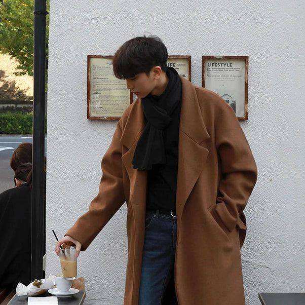 Sobretudo masculino Zellum longo de lã marrom, com cinto e 2 botões, sobre gola alta e jeans, tomando café em ambiente externo de cafeteria.