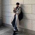 Sobretudo masculino Zellum longo bege liso, de algodão e poliéster, sobre gola alta preta e jeans, encostado em parede urbana usando celular, estilo casual-chique.