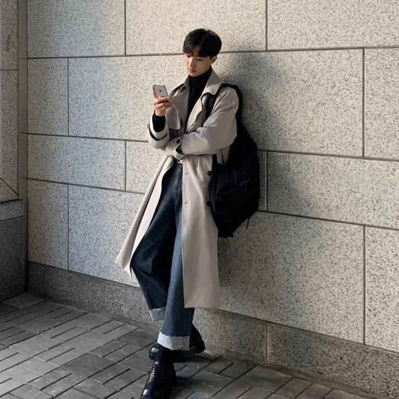 Sobretudo masculino Zellum longo bege liso, de algodão e poliéster, sobre gola alta preta e jeans, encostado em parede urbana, estilo moderno.