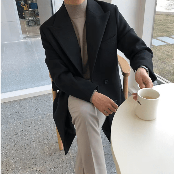 Sobretudo masculino Zellum Longo Mid preto, sobre gola alta bege e calça clara, sentado em cadeira de madeira em cafeteria, segurando xícara, evocando luxo.