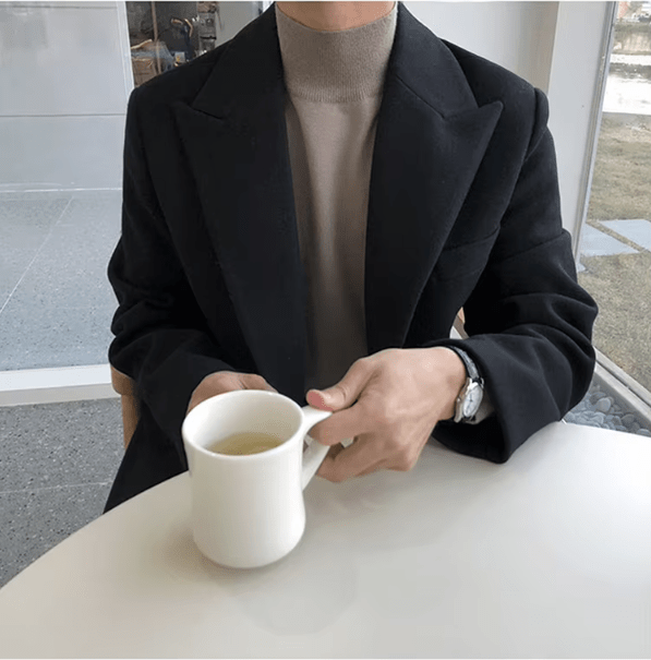 Sobretudo masculino Zellum Longo Mid preto, sobre gola alta bege, sentado em mesa, segurando xícara, evocando sofisticação em café.