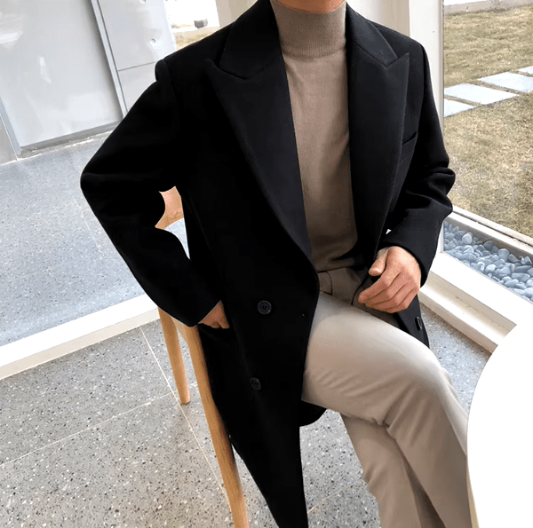Sobretudo masculino Zellum Longo Mid preto, sobre gola alta bege e calça clara, sentado em cadeira de madeira perto da janela, evocando sofisticação.