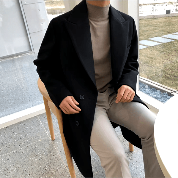 Sobretudo masculino Zellum Longo Mid preto, fechamento duplo, sobre gola alta bege e calça clara, sentado junto à janela, evocando sofisticação e conforto.