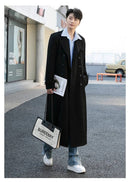Homem em trench coat preto longo, camisa listrada e jeans, segurando bolsa e revista em cenário urbano