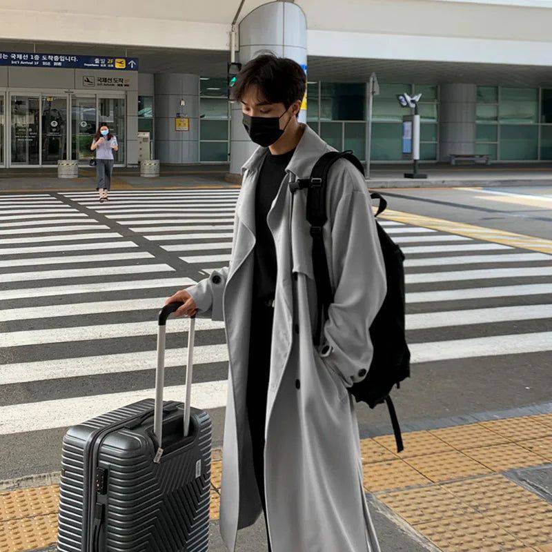 Sobretudo masculino Zellum longo e solto cinza claro, em frente ao aeroporto com mala e mochila, estilo casual-chique para viagem.