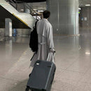 Homem de costas usando trench coat cinza claro, mala de viagem e mochila em terminal de aeroporto